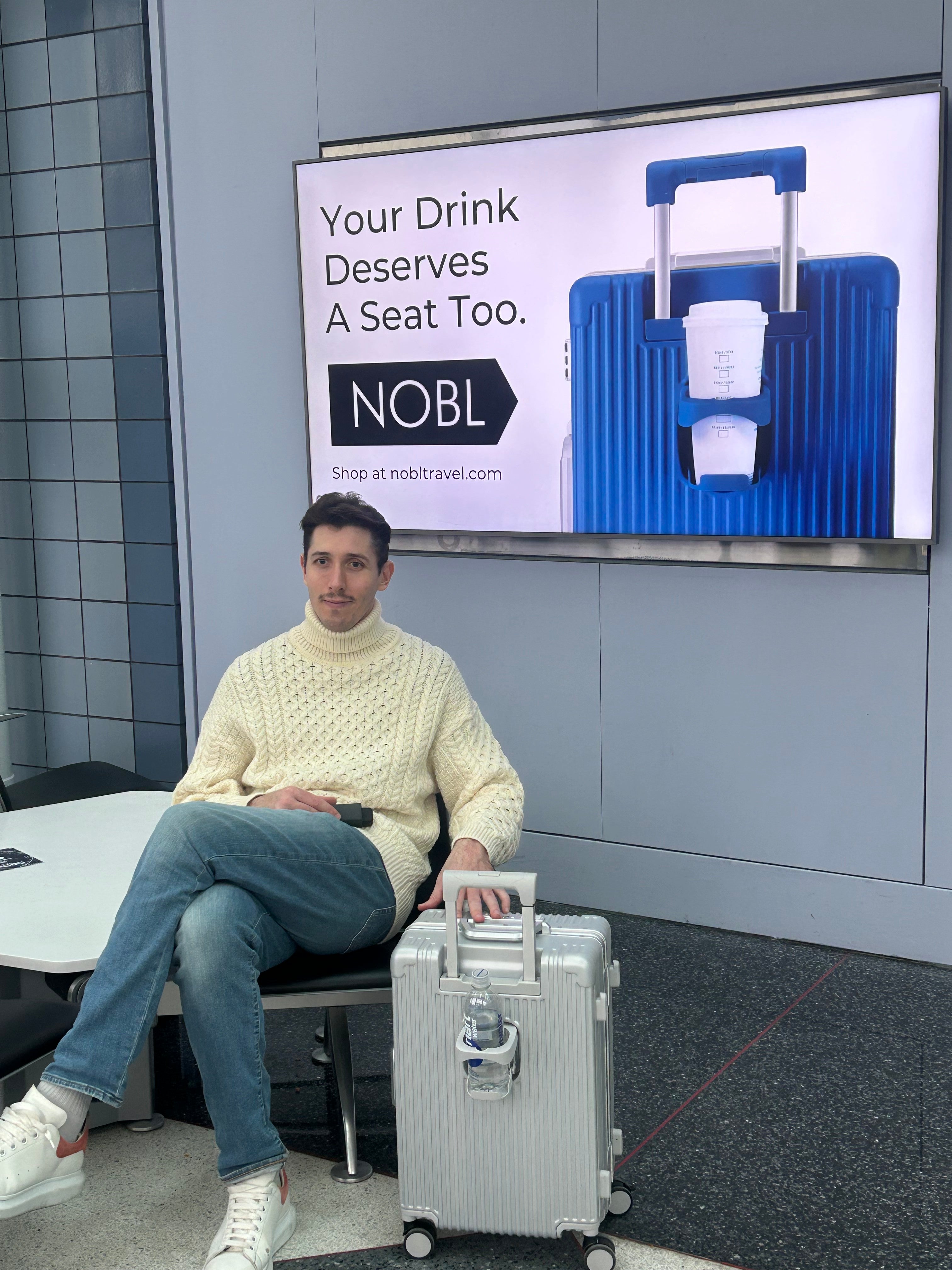 Traveler seated calmly at the airport beside a silver NOBL carry-on and NOBL billboard