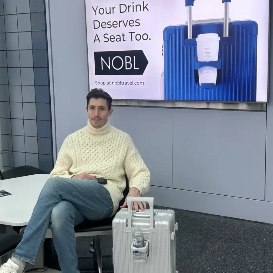 Traveler seated at the airport beside a silver NOBL carry-on with a cup holder